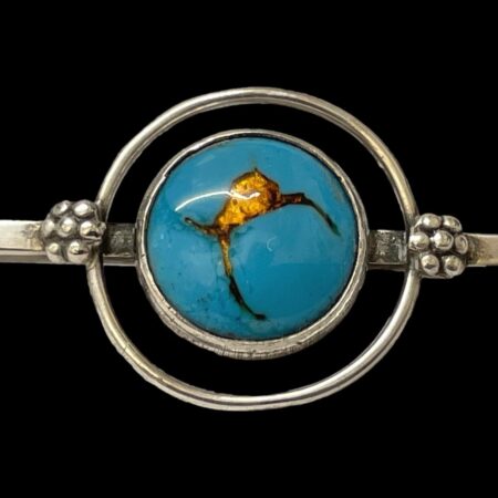 Main photo of Bernard Instone silver brooch seen from the front and focusing on the blue and gold enamel ball area at centre of the bar. The blue enamel circle to the centre has a fine silver ring around it. The blue enamel is of a dark turquoise colour with fissure like lines and a small crack opening which are all gold filled and looking like gold molten lava. There is a second 'satellite' ring of silver around the central blue circle adding another dimension.