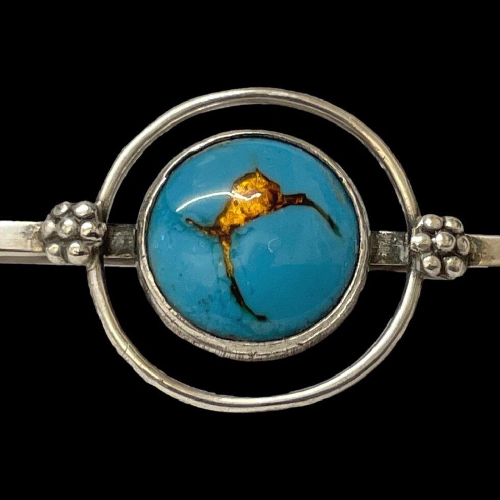 Main photo of Bernard Instone silver brooch seen from the front and focusing on the blue and gold enamel ball area at centre of the bar. The blue enamel circle to the centre has a fine silver ring around it. The blue enamel is of a dark turquoise colour with fissure like lines and a small crack opening which are all gold filled and looking like gold molten lava. There is a second 'satellite' ring of silver around the central blue circle adding another dimension.