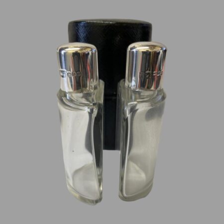 Main photo of Deco silver topped bottles displayed outside of their fitted leather case and placed in the foreground back to back. The bottles have a flat back and curved front to fit in the circular case. Each bottle has its own interior slot to keep them secure. The hallmark is visible on the lid of both bottles.