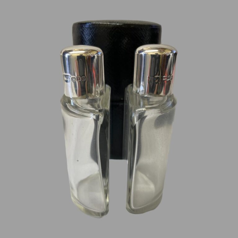 Main photo of Deco silver topped bottles displayed outside of their fitted leather case and placed in the foreground back to back. The bottles have a flat back and curved front to fit in the circular case. Each bottle has its own interior slot to keep them secure. The hallmark is visible on the lid of both bottles.