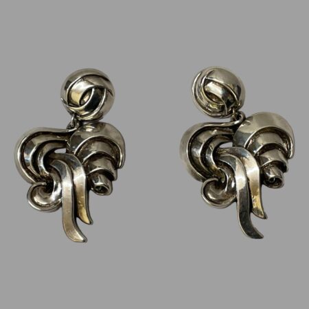 Main photo of both 1980s designer clip-ons displayed side by side on a flat surface and seen from the front. Earrings have a round section at the top where the clips are housed then almost like a swirled pattern of silver ribbon articulating below.