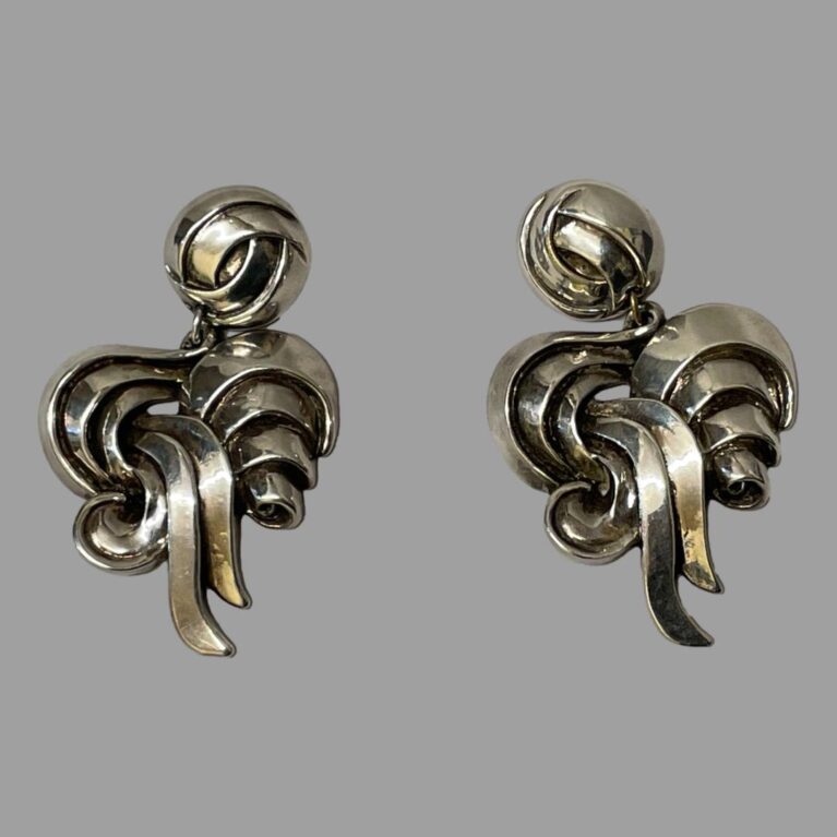 Main photo of both 1980s designer clip-ons displayed side by side on a flat surface and seen from the front. Earrings have a round section at the top where the clips are housed then almost like a swirled pattern of silver ribbon articulating below.