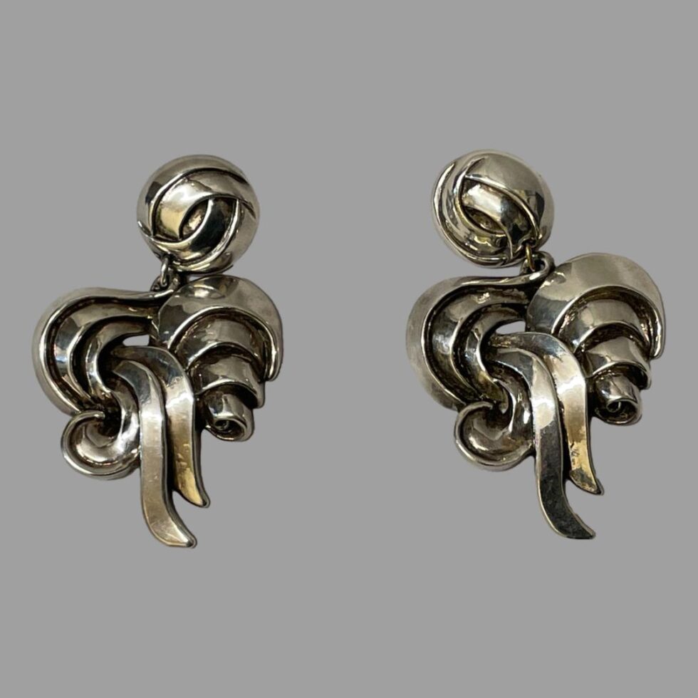 Main photo of both 1980s designer clip-ons displayed side by side on a flat surface and seen from the front. Earrings have a round section at the top where the clips are housed then almost like a swirled pattern of silver ribbon articulating below.
