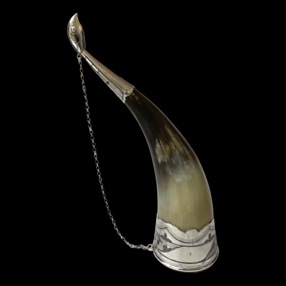 Photo of horn drinking vessel shown upright on its opening. The bird face and neck is at the top left with the chain hanging down from behind its neck which attaches to the silver rim around the vessel mouth.