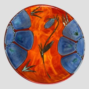 Main photo of Poole Himalayan poppy charger shown under a studio light and looking straight down from above. On the left and right of charger are 3 petals of half a blue poppy and to the centre a poppy bud that is waiting to bloom. The background colour is a flaming poppy red. A very striking statement piece from the famous Poole potteries.