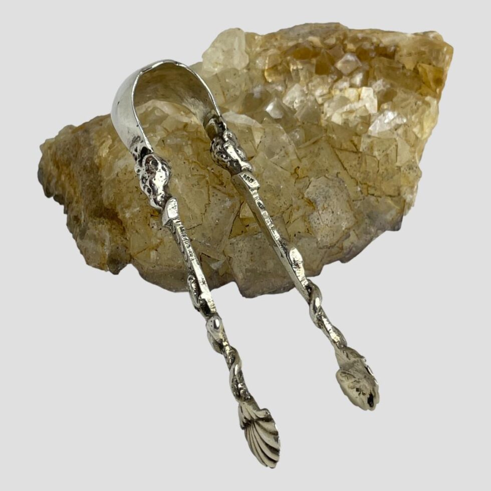 Photo of Continental silver sugar tongs displayed leaning against a rock of natural river quartz. At this angle, it is difficult to see the elaborate detailed design to the sides of the tongs.