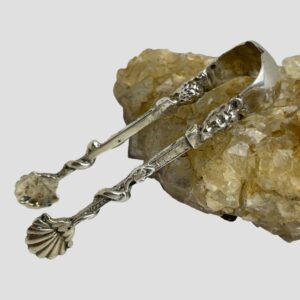 Main photo of Continental silver sugar tongs displayed leaning against a rock of river quartz and displayed with one side of the tongs in the foreground. Both sides are decorated the same with scallop spoon ends, a smooth snake entwined and crawling halfway up the tongs and finishing at the devil like heads at the top. The top of the tongs is plain silver.