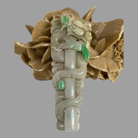 Main photo of long jade dragon pendant displayed with the wider top leaning against a rock of desert sand rose. The dragon is shown entwined around a thick bamboo pole as if ascending. Its head is looking straight up as it ascends reaching for the celestial heavens perhaps. The dragon has criss-cross scales throughout its body except for the head. The 3 small apple green jade inclusions are spaced out along the length of the long pendant.