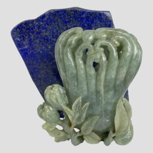 Main photo of jadeite calligraphy water pot displayed in front of a rock of lapis lazuli. The pot is a pale celadon green colour and contrasts well with the blue of the lapis. The side of pot on display has more flowers and foliage at the base. (2 partially open flowers to the left with leaves all along. The main body of the pot is like a group of bracken fiddleheads or croziers - shoots with a coiled tip like a shepherds crook. The shoots are joined at the base of the pot and separate to individual coiled shoots at top.