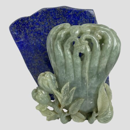 Main photo of jadeite calligraphy water pot displayed in front of a rock of lapis lazuli. The pot is a pale celadon green colour and contrasts well with the blue of the lapis. The side of pot on display has more flowers and foliage at the base. (2 partially open flowers to the left with leaves all along. The main body of the pot is like a group of bracken fiddleheads or croziers - shoots with a coiled tip like a shepherds crook. The shoots are joined at the base of the pot and separate to individual coiled shoots at top.
