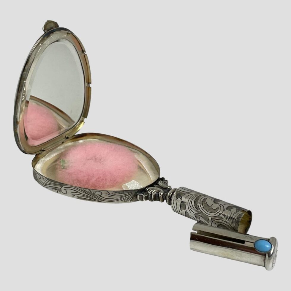 Photo of vanity mirror displayed diagonally across the page and open with interior mirror to top left corner and handle with lipstick holder removed in bottom right corner. The pink powder puff is inside the compact area.