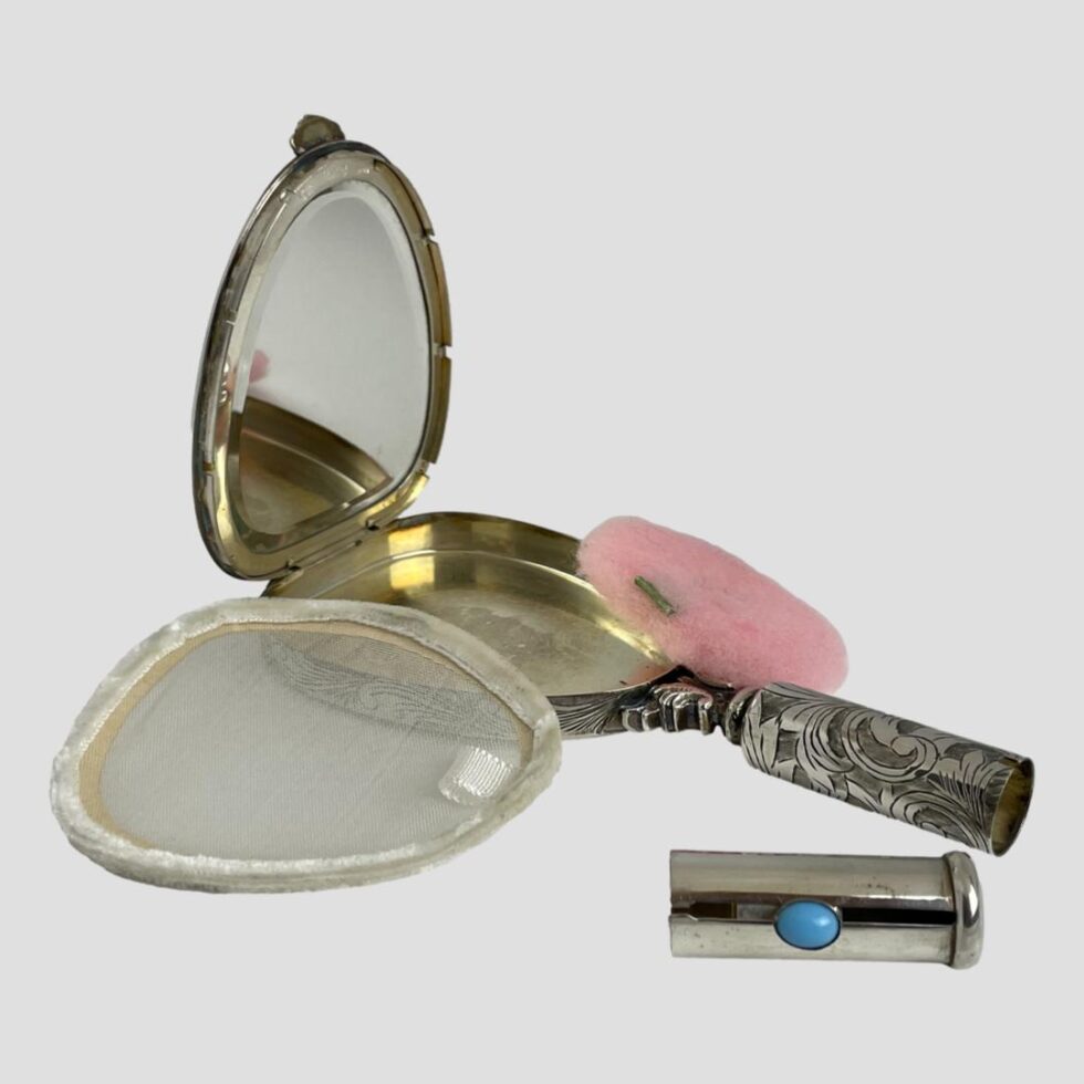 Photo of vanity mirror with all pieces removed. The mirror is laid diagonally across the page with the handle in the bottom right corner. The lip stick holder is jut in front of the handle, pink powder puff is in the background and leaning against the compact area while the net that keeps powder in place is leaning against the compact area in the foreground.