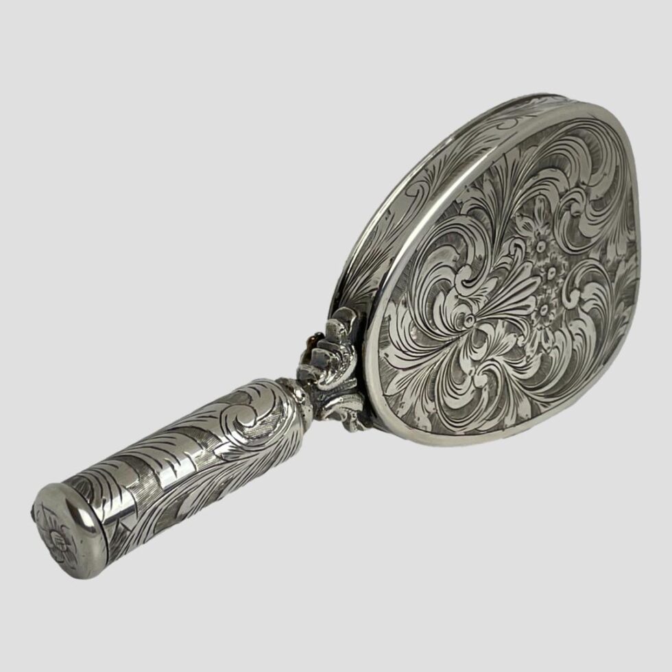 Photo of the vanity displayed upright on its side with the mirror side facing away. The mirror is displayed diagonally across the page with the handle in the bottom left corner. This side of the mirror is very elaborately embossed throughout including the sides of the mirror.