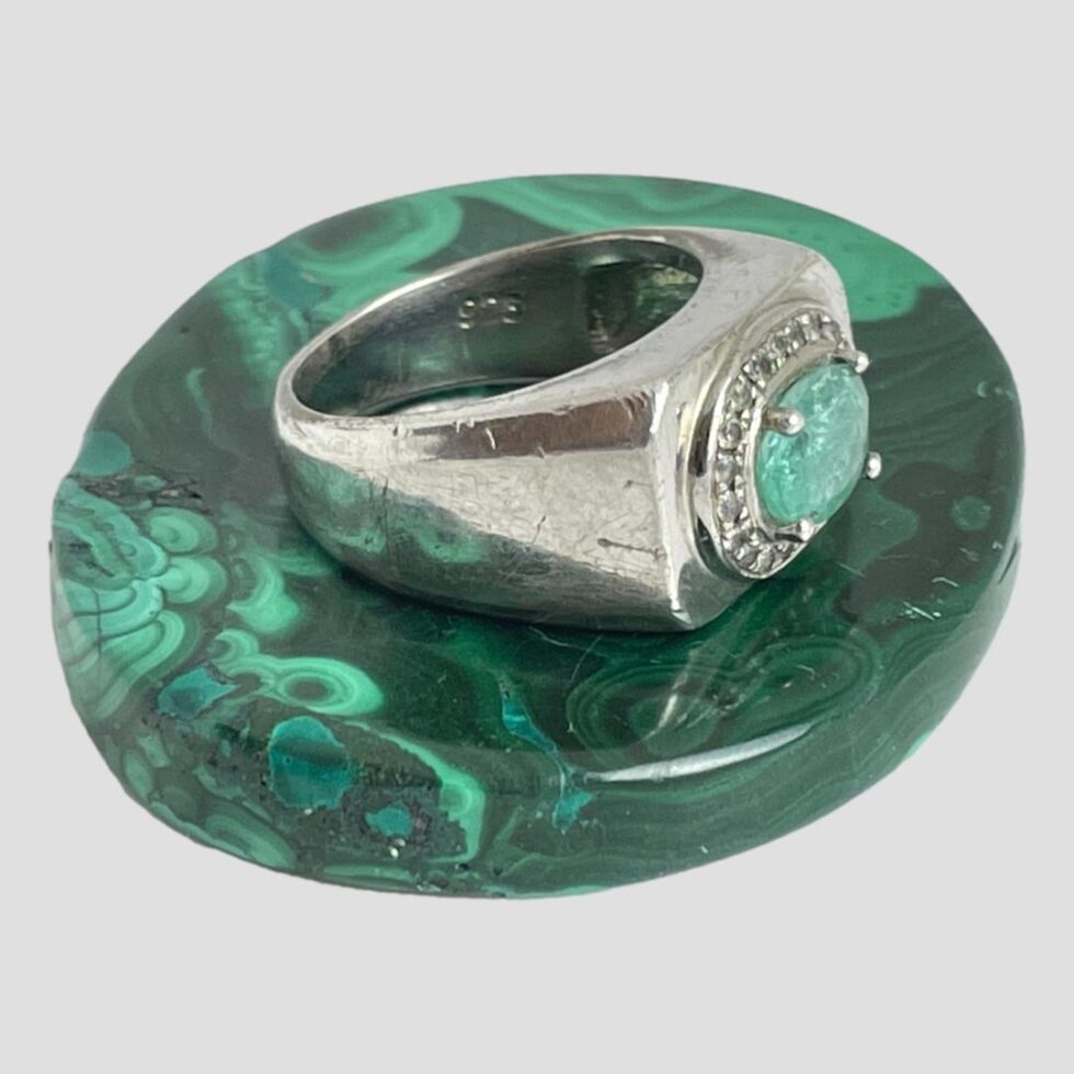 Photo of silver emerald ring still displayed on an oval malachite tablet with the ring front facing towards the bottom right corner. The shoulders of the ring are the same width as the ring front tapering down at the back but still thick.