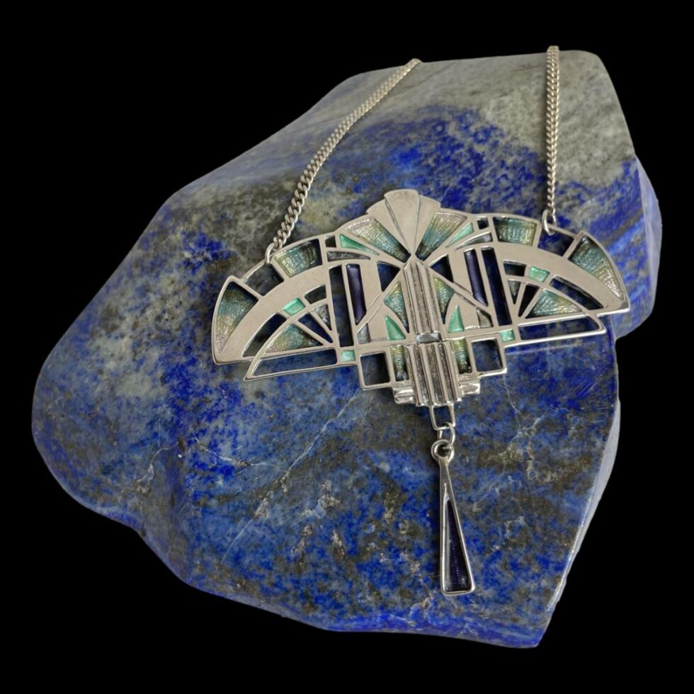 Main photo of Deco style silver necklace displayed with the pendant area resting on a rock of polished lapis lazuli and forward facing. The 'pendant' area is wide and would sit across the front of the collar bone. The lovely geometry of Art Deco design and the colours of the era - a little hint of suffragettes perhaps? There is a small thin teardrop shaped piece hanging down from the centre of the pendant. A very striking statement piece!