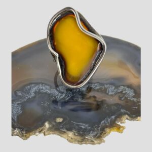 Main photo of large Baltic amber ring displayed on top of a slice of natural agate with the ring front forward facing. The egg yolk amber is a beautiful rich yellow with orange hues. The ring is shaped a little liked the continent of Africa at a stretch of the imagination! The egg yolk amber has a ring or frame of cognac amber all around it. The silver setting is very abstract with parts of the silver frame intruding over the amber which adds extra wow factor to this large statement ring.