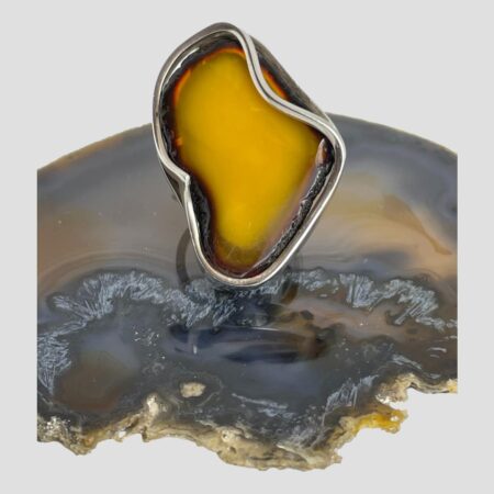 Main photo of large Baltic amber ring displayed on top of a slice of natural agate with the ring front forward facing. The egg yolk amber is a beautiful rich yellow with orange hues. The ring is shaped a little liked the continent of Africa at a stretch of the imagination! The egg yolk amber has a ring or frame of cognac amber all around it. The silver setting is very abstract with parts of the silver frame intruding over the amber which adds extra wow factor to this large statement ring.