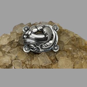 Main photo of Georg Jensen dove brooch displayed on a rock of natural river quartz with the brooch front forward facing. The dove is central to the brooch with body facing right but head bent back preening its wing. The brooch has rounded corners with a ball to centre and there are some foliage decorations around the dove.