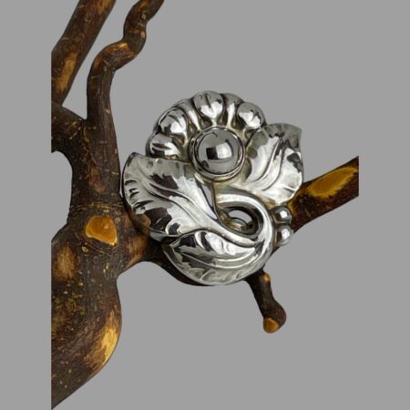 Main photo of Georg Jensen floral brooch displayed perched on a dried cherry tree twig with the brooch front forward facing. The design has 3 large leaves at the base with just under half a flower sticking out from behind them. The flower has a silver ball centre with small petals - a little sunflower like.