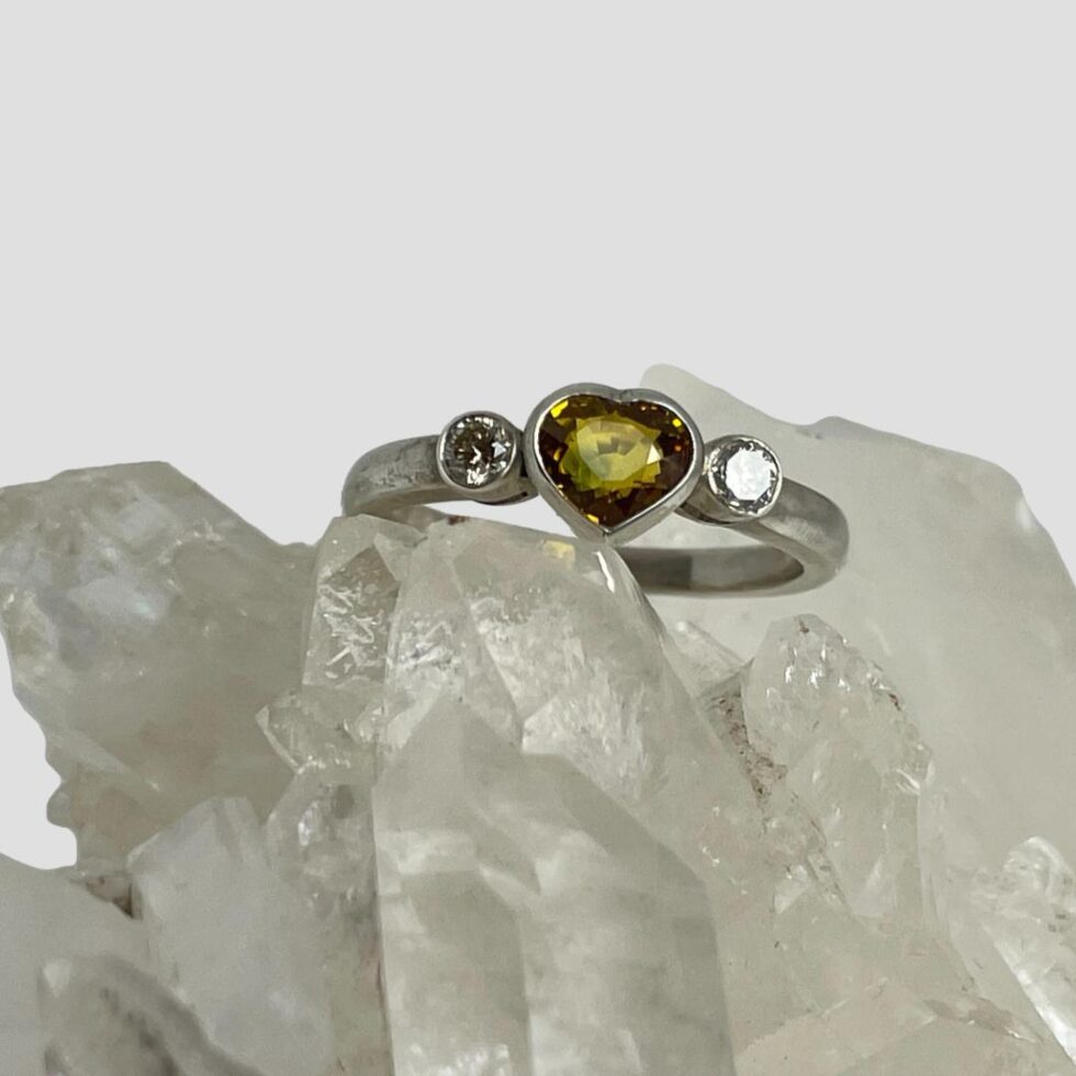 Main photo of yellow sapphire diamond ring displayed on a rock of natural quartz crystal with the ring front forward facing. The heart shaped yellow sapphire has a very slight orange hue and has been beautifully faceted. The round cut diamonds to either side of the heart sapphire are a generous size and sparkle beautifully.