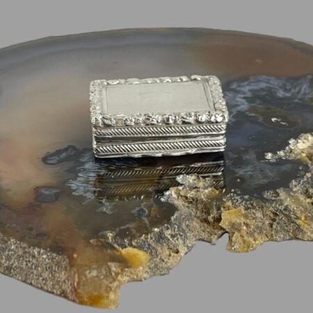 Main photo of Edward VII silver vinaigrette displayed on top of a slice of natural agate and displayed with the opening front side in the centre foreground. This vinaigrette is incredibly small but given its size, there is an incredible amount of detail in the design. Engine turned background with rope effect at the sides and a pretty floral detail all around the top edge. There is also a tiny empty cartouche on the top of the lid!