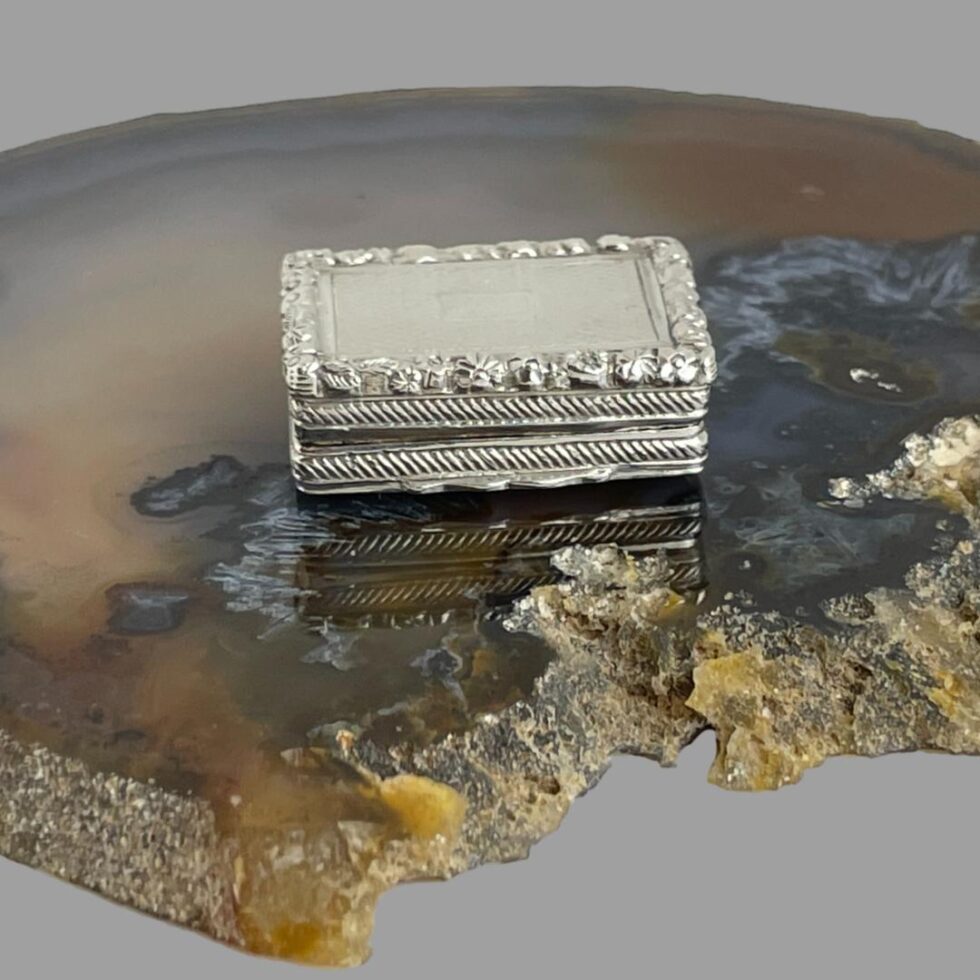 Main photo of Edward VII silver vinaigrette displayed on top of a slice of natural agate and displayed with the opening front side in the centre foreground. This vinaigrette is incredibly small but given its size, there is an incredible amount of detail in the design. Engine turned background with rope effect at the sides and a pretty floral detail all around the top edge. There is also a tiny empty cartouche on the top of the lid!