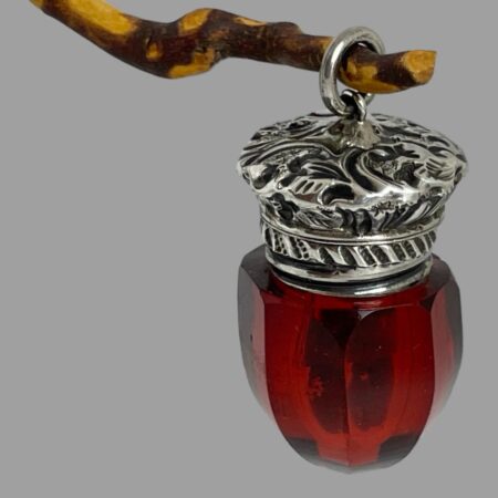 Main photo of antique smelling salts bottle displayed hanging from a wooden twig by its ring bail. The octagonal shaped glass bottle is crafted to look like an acorn with the bottom coming to a point. The colour of the glass is a deep rich red. The hinged top is heavily engraved.