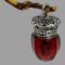 Main photo of antique smelling salts bottle displayed hanging from a wooden twig by its ring bail. The octagonal shaped glass bottle is crafted to look like an acorn with the bottom coming to a point. The colour of the glass is a deep rich red. The hinged top is heavily engraved.