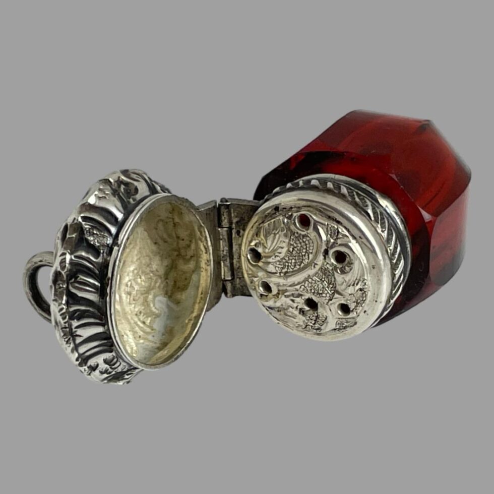 Photo of antique smelling salts bottle displayed laid on a flat surface with the hinged top open revealing the perforated top. There are 6 small holes in the top which are not uniform in spacing.