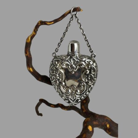 Main photo of silver heart scent bottle displayed hanging from a twig by its chain. One side of the heart is on display. Embossed detail covers most of the heart with just the screw top and cartouche in smooth silver.
