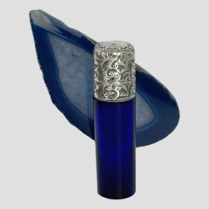 Main photo of antique portable scent bottle displayed stood upright in front of a slice of blue agate. The opening front side of the bottle is in the foreground. The blue bottle is small, cylindrical and easily portable in a bag or even pocket. It has a heavily engraved hinged top which makes up about a third of the entire bottle.