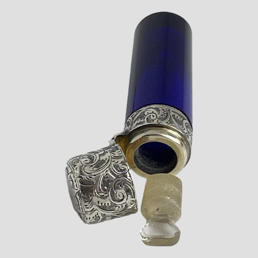 Photo of antique scent bottle displayed on its side with the lid shown open and glass stopper removed and placed in front of the bottle opening.