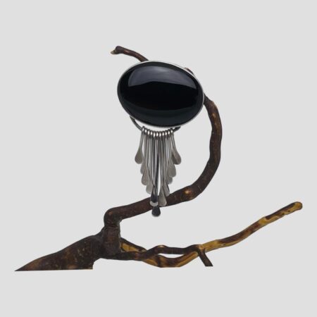 Main photo of Mexican onyx brooch pendant shown hanging from a twig of dried cherry and displayed with the brooch front forward facing. The large oval onyx is horizontally long with graduating silver strands with teardrop ends hanging below from a curved bar underneath the stone. The longest of these strands is at the centre and the strands graduate shorter on both sides. They are reminiscent of suns rays