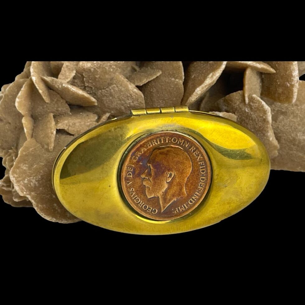 Main photo of antique brass snuff/twist box displayed upright with the copper George V penny inlaid top on display. The box is leaning against a rock of desert sand rose in the background. The brass body with the copper coin gives an appearance of an eye. Very shiny metal piece in very good order.