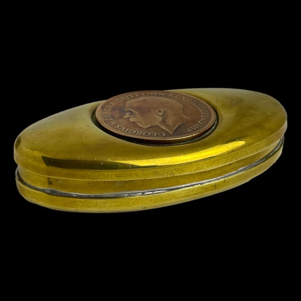 Photo of miners brass snuff/twist box laid on a flat surface at a diagonal angle with the opening edge side facing towards the bottom right corner.