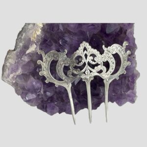 Main photo of Edwardian silver hair comb displayed upright and leaning against a natural rock of amethyst. The comb front is forward facing This antique item has stood the test of time however, it is very fine silver and quite pliable so extra care is needed when handling. There are 3 prongs. The 2 on either end have a stylised stem with leaf like protrusions at the sides and a single small flower at the curved tip. The central prong housed the most decoration with 2 stylised stems splitting out and an elaborate and intricate central area made up of more swirls. All of the silver at the top of the comb has engraved detail. A fabulous antique piece which may not be relevant for use today although would not look amiss on a flamenco dancer.