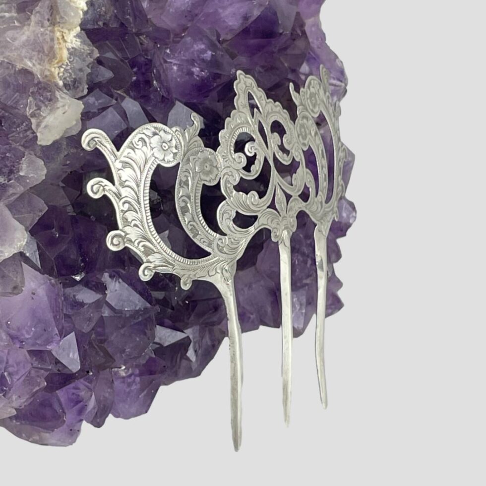 Photo of Edwardian silver hair comb still displayed upright and leaning against a natural rock of amethyst this time from a side angle with comb front facing to the bottom right corner.