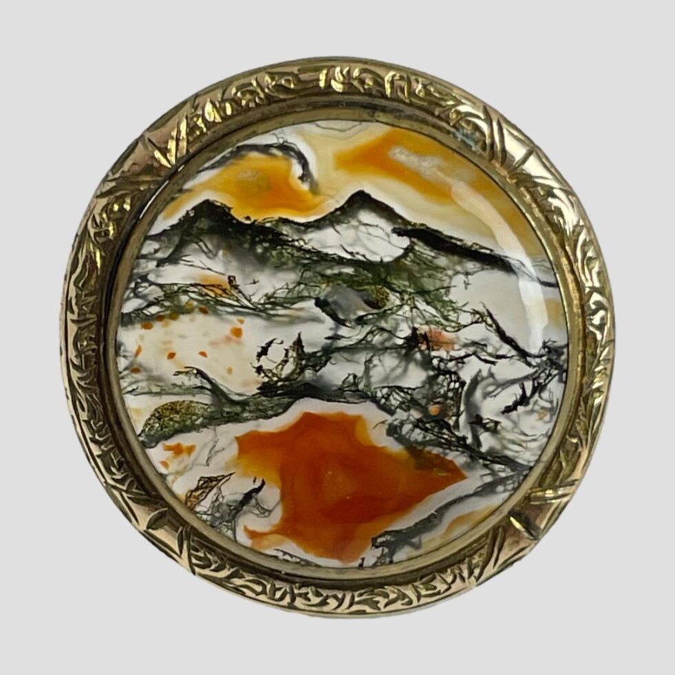 Photo of moss agate brooch this time displayed on a plain light grey surface with the brooch front forward facing in close up. The image of mountains, valleys and lake is very noticeable. The gilt frame has a simple notch work decoration and compliments the colours of the moss agate which is in clear, clack, orange and terracotta colours.