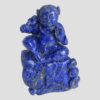 Main photo of vintage carved lapis figural sculpture with its front forward facing. The figure, a young child like male is sat on a rock being lapped by the sea with a large fish possibly a carp, leaping from the water to the bottom left. The boy has a raised left hand (right in the photo) with palm forward facing and his right arm is holding a ring from which a piece of cloth is attached. The boys hair is bunched up on the sides into tight rings and he wears a neck band. Believed to be a good luck ornament.
