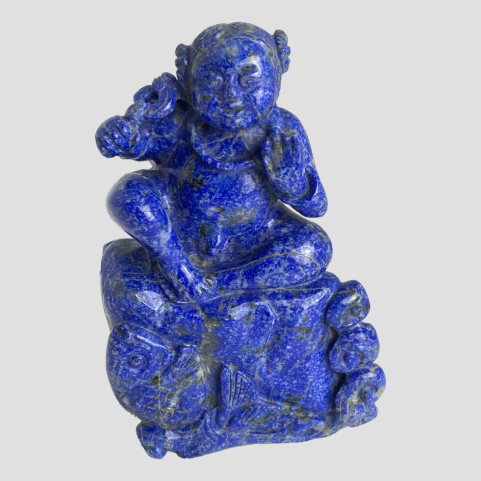 Main photo of vintage carved lapis figural sculpture with its front forward facing. The figure, a young child like male is sat on a rock being lapped by the sea with a large fish possibly a carp, leaping from the water to the bottom left. The boy has a raised left hand (right in the photo) with palm forward facing and his right arm is holding a ring from which a piece of cloth is attached. The boys hair is bunched up on the sides into tight rings and he wears a neck band. Believed to be a good luck ornament.