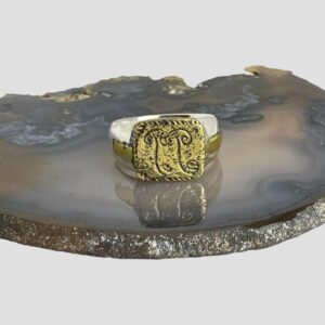 Main photo of upcycled floral Medieval ring displayed on top of a slice of agate with the ring front facing forward. The Medieval fragment has a rectangular section which forms the ring front with 2 stylised floral images engraved in the centre. These could even be bramble or mushrooms. Lines have been scored around the rectangle edge. A short alloy section makes part of one shoulder and a longer strip of alloy extends all the way round to the back of the ring. These strips are set within the chunky silver band and have no decoration just plain gilt coloured metal. How wonderful to be able to wear an archaeological find on your finger!