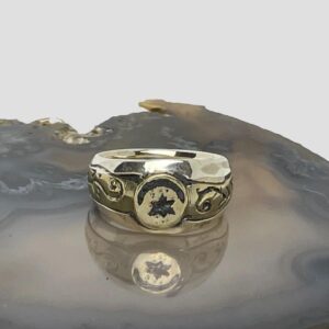 Main photo of upcycled Medieval fragment ring displayed on top of a slice of natural agate with the ring front facing forward. The crescent moon is set across the top of the circular bronze centre with the star underneath it. The bronze fragment extends on either side to make the shoulders. These areas have some embossed detail of possibly vines. The band is in solid sterling silver and the plainness of it compliments the Medieval bronze fragment perfectly.