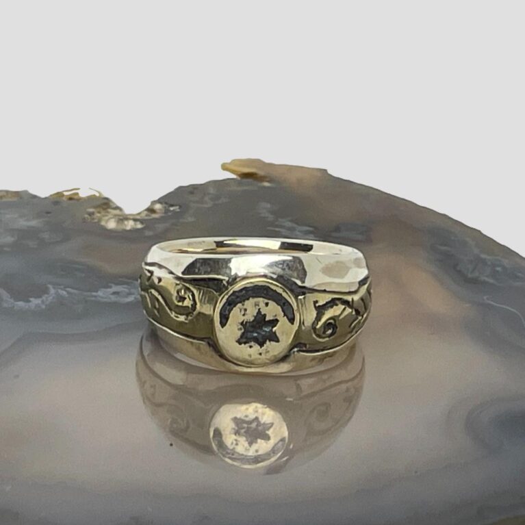 Main photo of upcycled Medieval fragment ring displayed on top of a slice of natural agate with the ring front facing forward. The crescent moon is set across the top of the circular bronze centre with the star underneath it. The bronze fragment extends on either side to make the shoulders. These areas have some embossed detail of possibly vines. The band is in solid sterling silver and the plainness of it compliments the Medieval bronze fragment perfectly.