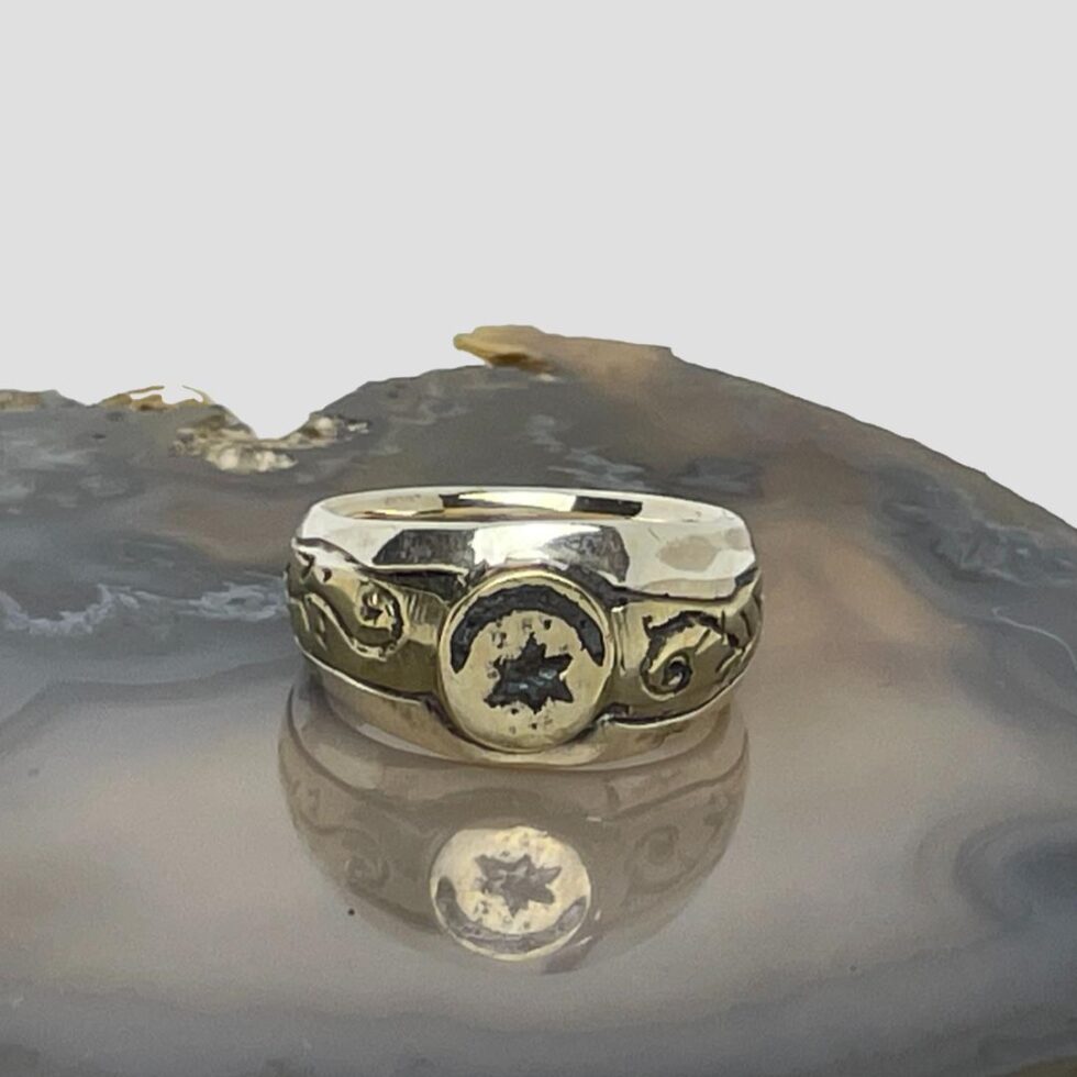 Main photo of upcycled Medieval fragment ring displayed on top of a slice of natural agate with the ring front facing forward. The crescent moon is set across the top of the circular bronze centre with the star underneath it. The bronze fragment extends on either side to make the shoulders. These areas have some embossed detail of possibly vines. The band is in solid sterling silver and the plainness of it compliments the Medieval bronze fragment perfectly.