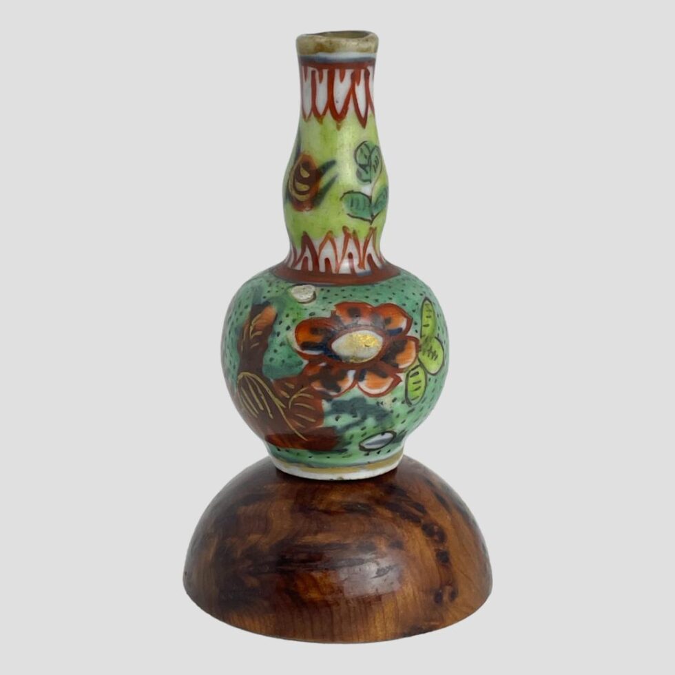 Main photo of Kangxi miniature gourd vase displayed on top of a small wooden display dome. The vase has a base colour of a bluey green on the lower bulbous area and a bright light leafy green on the neck The flowers range from orangey red to deep red with golden accents and a couple of random small white circles. The blue green of the bulbous area has black dots throughout. The neck is separated from the bulbous base by a red colour with small white triangles pointed upward all the way around. These are lined in red with a red flick at the centre. The bright light leaf green neck has a few minor floral decorations and the rim of the vase has downward facing triangles mirroring the triangles at waist. The dome stand is not included.