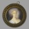 Main photo of antique French miniature portrait displayed upright with the portrait figure looking straight to camera. The ring for hanging the picture is seen off centre and to the right and would be ideal to get it reset. The picture is very securely in place though and may need a professional or at least someone who knows what they're doing to fix it. The lady is wearing a low cut top in pale baby blue with white lace trim. Her curly locks are short in the image and appear white blonde. A small posy with a blue flower sits in her hair which matches the colour of her piercing eyes. There is an evocative feel to this miniature which follows you around the room!
