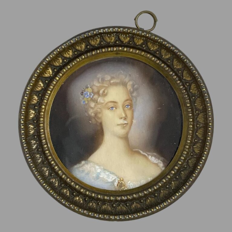 Main photo of antique French miniature portrait displayed upright with the portrait figure looking straight to camera. The ring for hanging the picture is seen off centre and to the right and would be ideal to get it reset. The picture is very securely in place though and may need a professional or at least someone who knows what they're doing to fix it. The lady is wearing a low cut top in pale baby blue with white lace trim. Her curly locks are short in the image and appear white blonde. A small posy with a blue flower sits in her hair which matches the colour of her piercing eyes. There is an evocative feel to this miniature which follows you around the room!