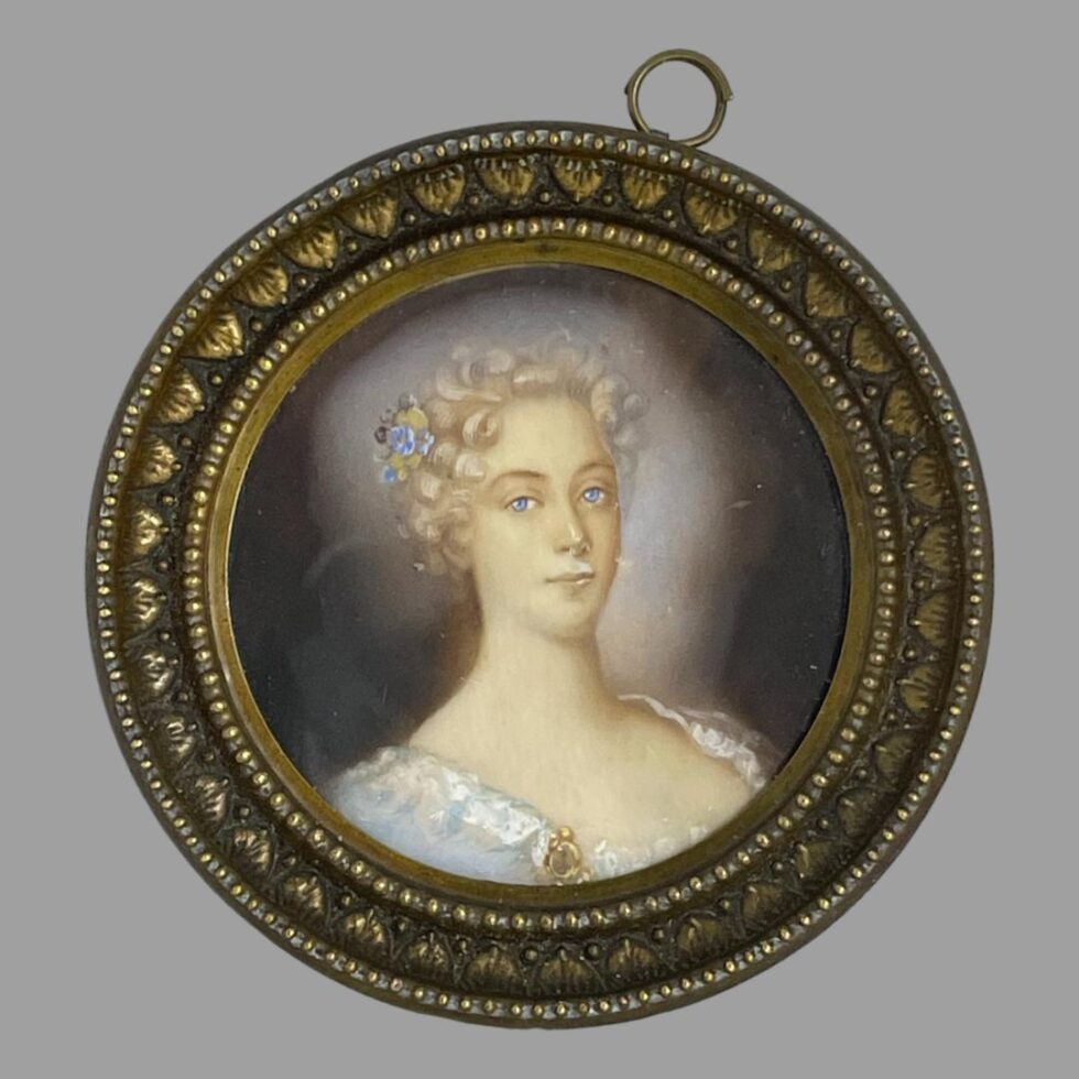 Main photo of antique French miniature portrait displayed upright with the portrait figure looking straight to camera. The ring for hanging the picture is seen off centre and to the right and would be ideal to get it reset. The picture is very securely in place though and may need a professional or at least someone who knows what they're doing to fix it. The lady is wearing a low cut top in pale baby blue with white lace trim. Her curly locks are short in the image and appear white blonde. A small posy with a blue flower sits in her hair which matches the colour of her piercing eyes. There is an evocative feel to this miniature which follows you around the room!