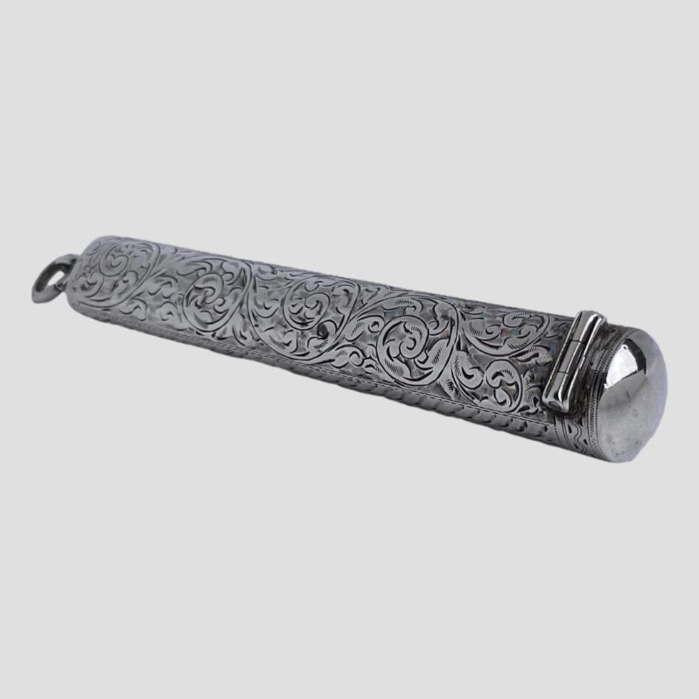 Photo of antique silver cheroot case displayed on a flat surface this time with the hinged opening end in the bottom left foreground with the embossed scrollwork covering the whole back.