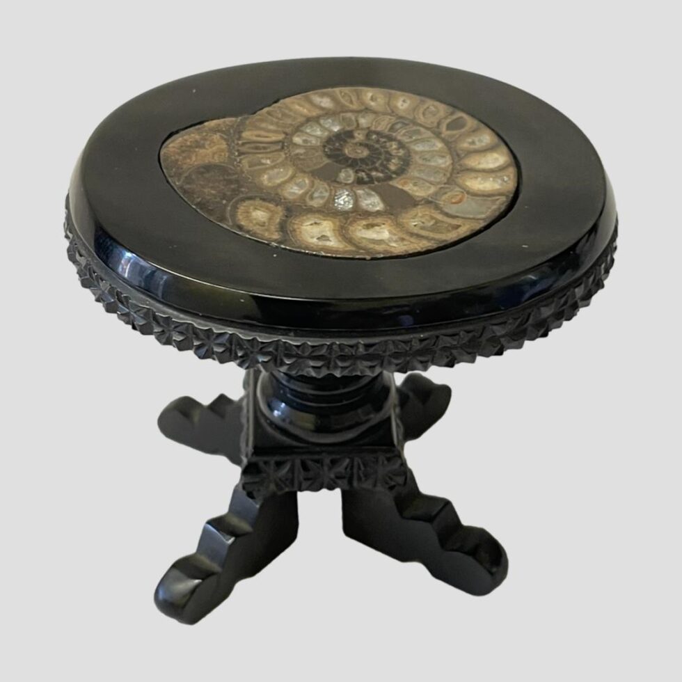 Main photo of Victorian jet miniature table displayed from slight height looking diagonally down at the ammonite in the table top. The jet has been expertly carved with detail to the outer table edge as well as above the table legs. The table is a pedestal variety with a central pillar and 4 feet. There are some signs of wear as well as old repairs. The ammonite in the table top is sectioned and in fine condition.